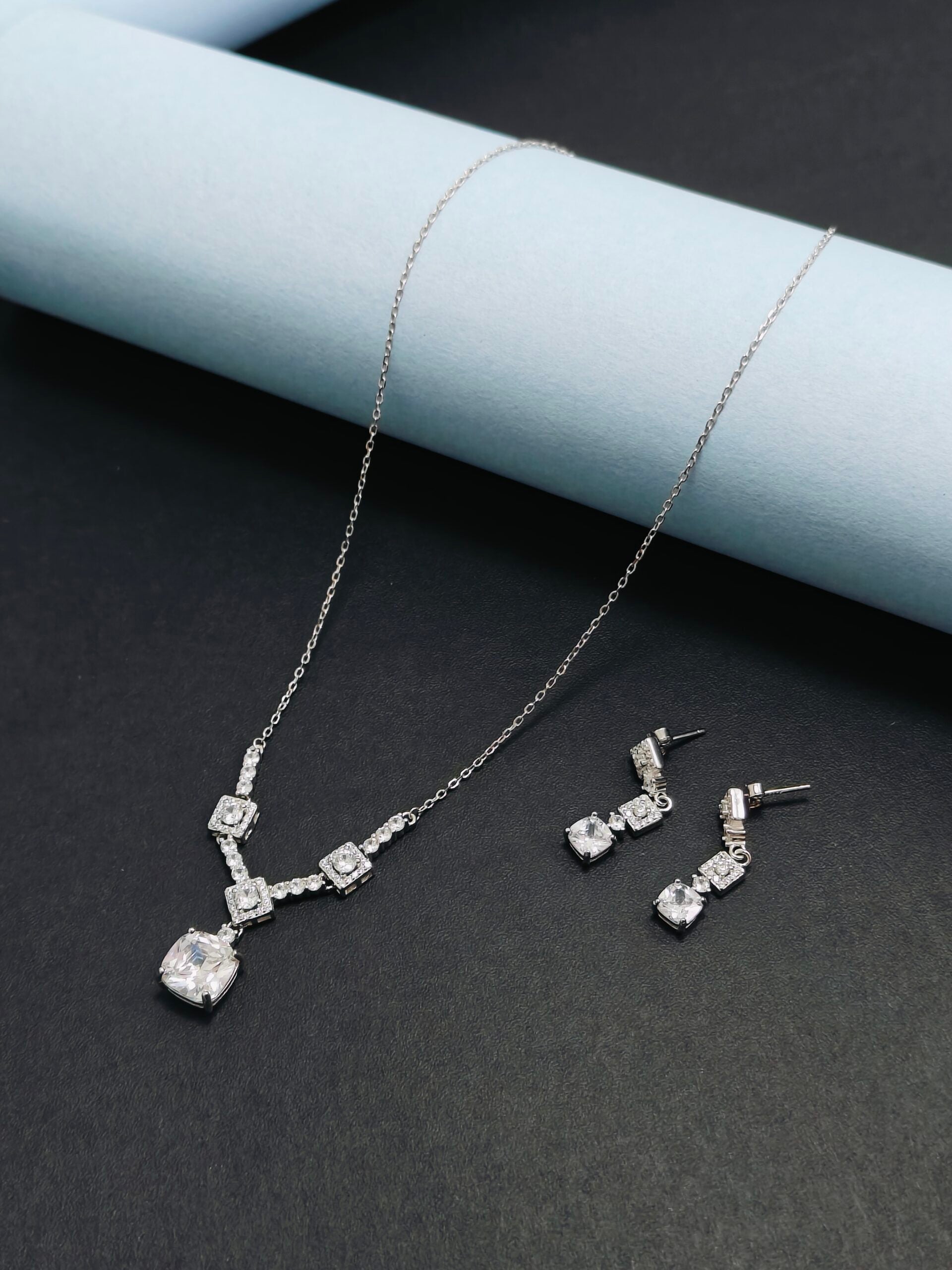 Silver necklace and earrings set on a dark surface with a light blue cylindrical object.